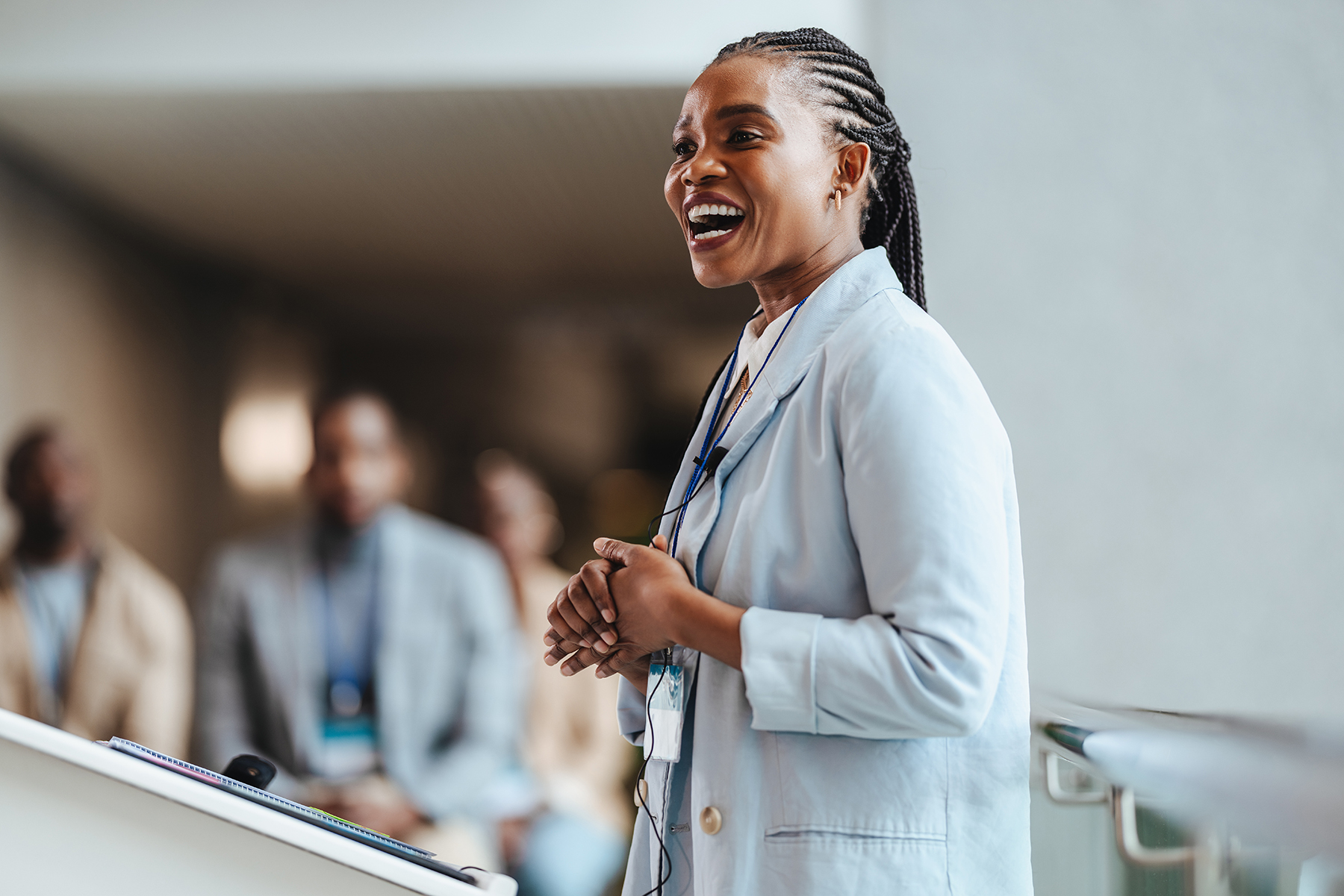 Professional African woman with a radiant smile engages attendees as she speaks at a business conference. With a blurred team in the backdrop, she embodies leadership and confidence.